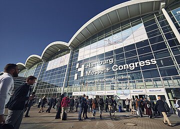 Blick auf den Eingang Mitte der Hamburg Messe und Congress mit zahlreichen Messebesuchenden vor der großen Glasfassade.