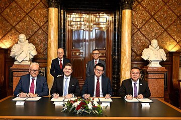 Fünf Personen sitzen an einem Tisch und unterzeichnen Dokumente, dahinter zwei stehende Personen, prunkvoller Raum mit Marmorsäulen und Büsten.