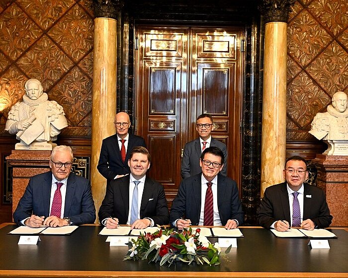 Fünf Personen sitzen an einem Tisch und unterzeichnen Dokumente, dahinter zwei stehende Personen, prunkvoller Raum mit Marmorsäulen und Büsten.