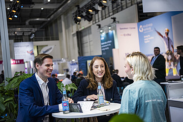 Drei Personen sitzen an einem Stehtisch auf einer Messe und führen ein Gespräch. Im Hintergrund sind Messestände, Besucherinnen und Besucher sowie Ausstellungsflächen in einer modernen Veranstaltungshalle zu sehen.
