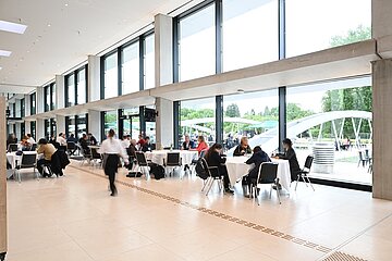 „Helle Lounge im CCH mit großen Fensterfronten, mehreren runden Tischen und Personen, die zusammensitzen.“