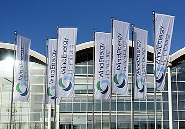 Fahnen mit dem Logo der WindEnergy Hamburg vor dem gläsernen Messegebäude bei strahlend blauem Himmel.