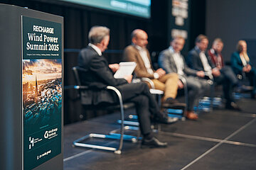 Podiumsdiskussion beim „Wind Power Summit 2025“ mit mehreren Fachleuten auf der Bühne, Fokus auf Veranstaltungsplakat im Vordergrund.
